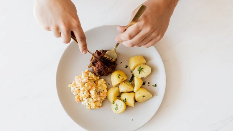 Top view of crop unrecognizable male with cutleries slicing delicious beef cutlet garnished with boiled potatoes and parsley and portion of Russian meat salad on plate placed on white table